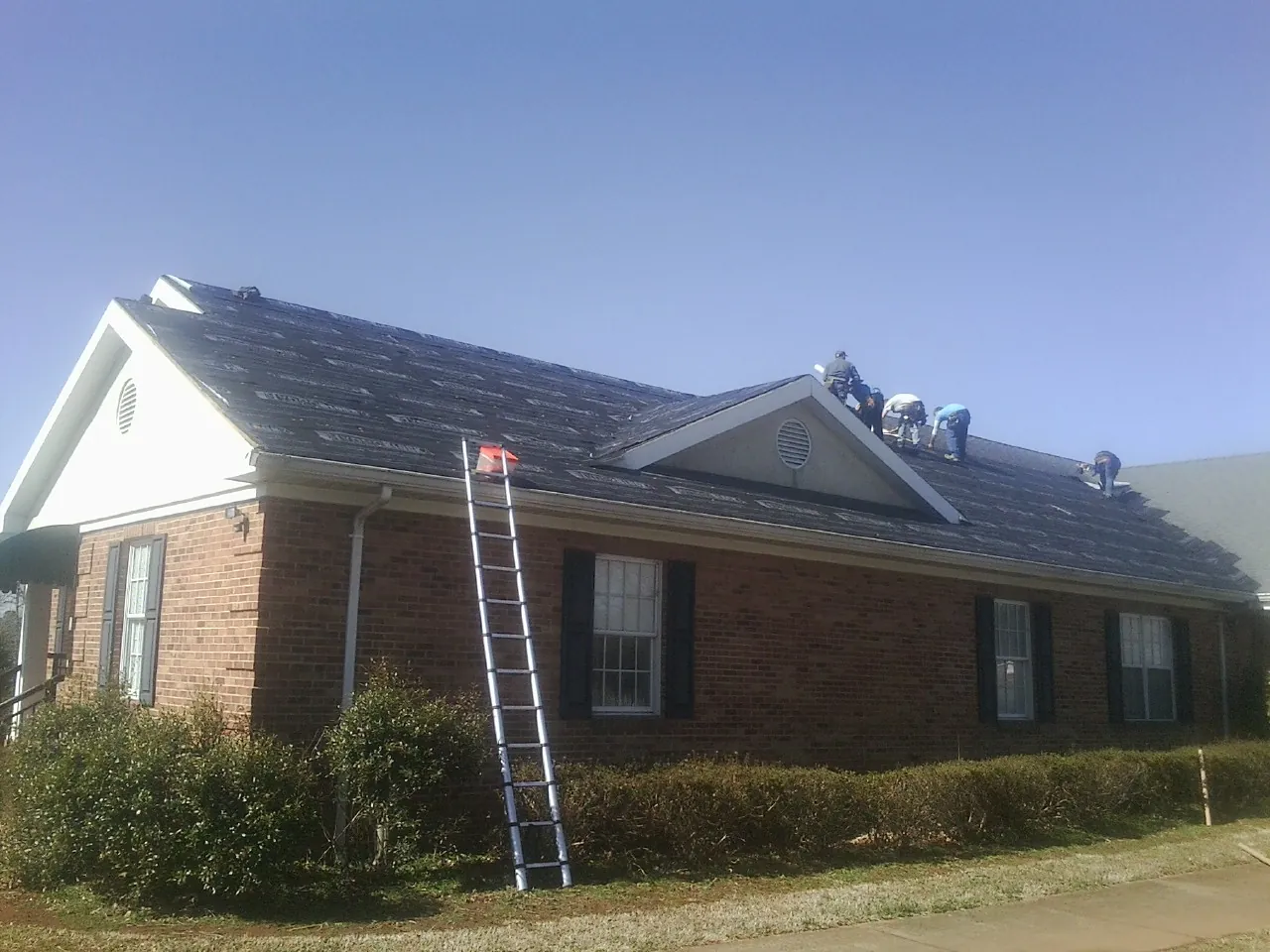 Crew tearing shingles off a brick ranch roof with a ladder set — Metal Roof Repair work by our crew in Oak Hill